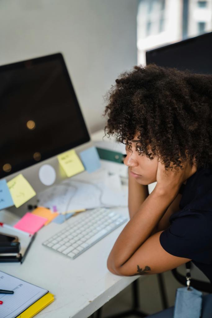 A black young woman seems exasperated with her hands on on either side and holding her head. She has her computer screen on the table where she is working remotely.