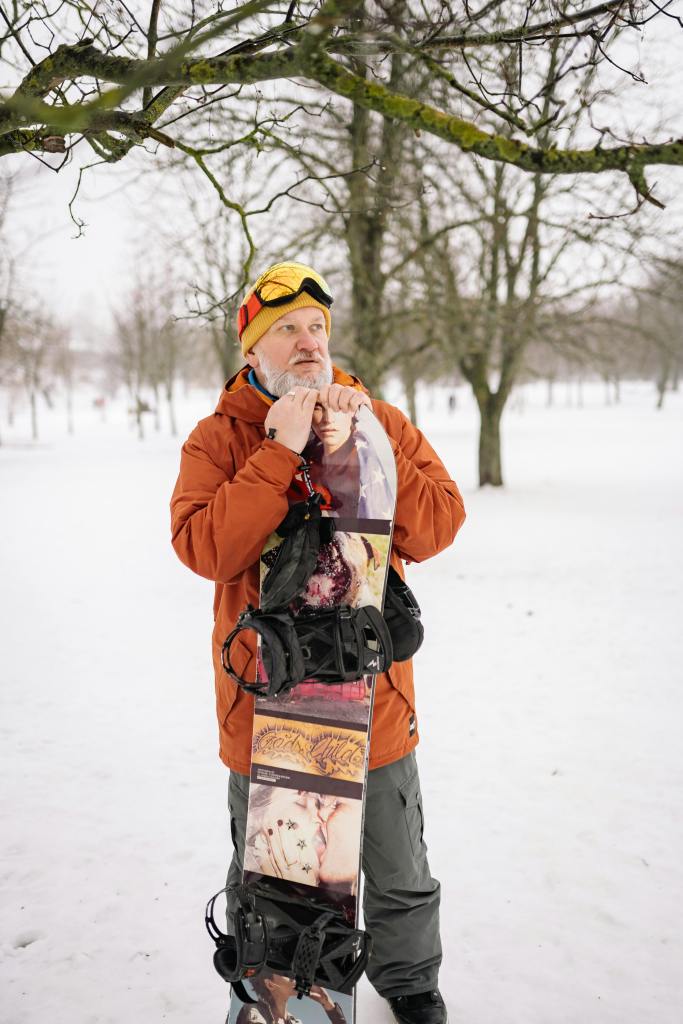 An elderly man standing and holding a skiing equipment. He is standing in an outdoor space with snow all around